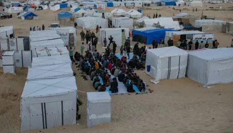 epa11244968 Displaced people sit together next to their tents to eat during the holy month of Ramadan at a collective Iftar meal organized by a youth volunteer group in a makeshift camp on the Egyptian border near Rafah, in the southern Gaza Strip, 26 March 2024. The Muslims' holy month of Ramadan is the ninth month in the Islamic calendar and it is believed that the revelation of the first verse in the Koran was during its last 10 nights. It is celebrated yearly by praying during the night time and abstaining from eating, drinking, and sexual acts during the period between sunrise and sunset. It is also a time for socializing, mainly in the evening after breaking the fast and a shift of all activities to late in the day in most countries. EPA/HAITHAM IMAD