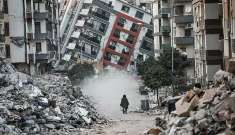 epaselect epa10473572 A woman walks in front of a collapsed building while demolition teams work after a powerful earthquake in Hatay, Turkey, 17 February 2023. Almost 44,000 people have died and thousands more are injured after two major earthquakes struck southern Turkey and northern Syria on 06 February. Authorities fear the death toll will keep climbing as rescuers look for survivors across the region. EPA/SEDAT SUNA