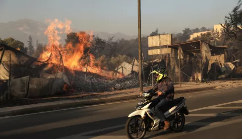 epa11546917 A person riding a scooter watches as a wildfire engulfs a firewood business, in Penteli, northeast of Athens, Greece, 12 August 2024. The wildfire that broke out in Varnavas on 11 August afternoon continued to rage in eastern Attica on 12 August, fanned and spread to a front extending more than 20 kilometers. According to the fire department, the fire-fighting effort is extremely difficult as the wind keeps changing direction, while the three main fronts of concern are in Grammatiko, Penteli and the Anatoli settlement in Nea Makri. EPA/GEORGE VITSARAS