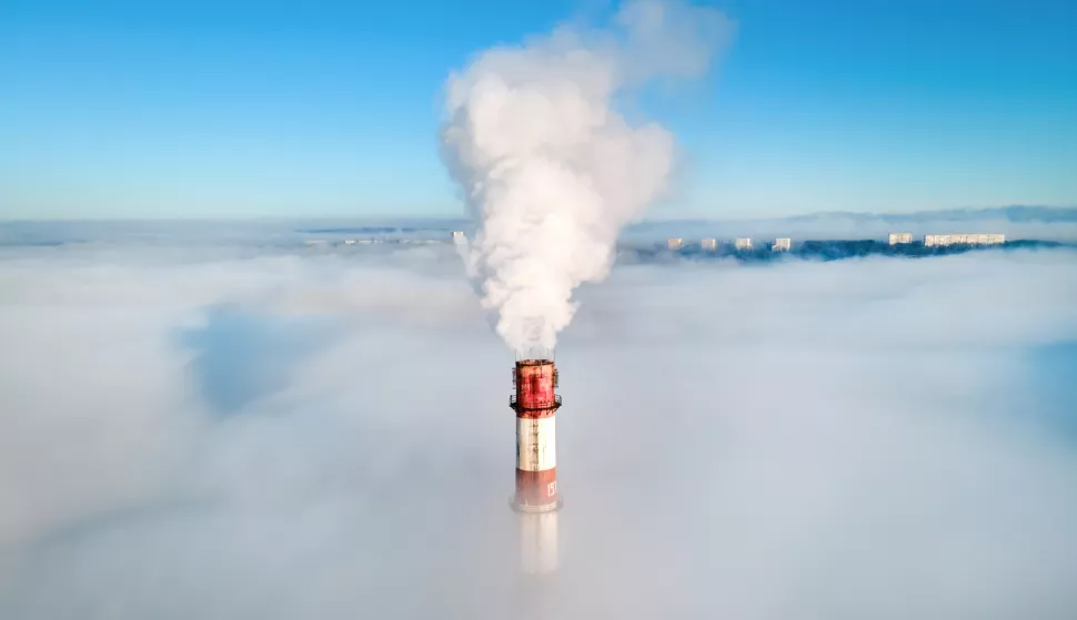 Aerial drone view of thermal station's tube visible above the clouds with smoke coming out. Buildings on the background. Blue and clear skyindustrija zagađenje fosilna goriva dim dimnjak freepik