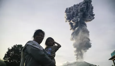 epa11826439 A woman carries a child as Mount Ibu spews volcanic material up to the sky in the background during the eruption in West Halmahera, North Maluku province, Indonesia, 15 January 2025. Indonesia's Vulcanology and Geological Disaster Mitigation Center (PVMBG) raised the alert level of the volcano to the highest following the eruption. EPA/STR