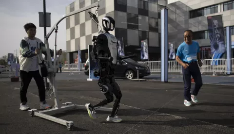 epa12036662 Zhang Xianghua (71), a runner who will take part in the race, joggs next to a humanoid robot ahead of a humans and robots race in Beijing, China, 17 April 2025. Beijing will host a half marathon on April 19, in which humans and robots will participate. EPA/ANDRES MARTINEZ CASARES