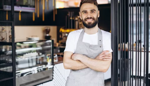 Male barista standing and smiling at a coffee houseKoliko sezonac može zaraditi?Istražili smo koliko traže radnici, a koliko nude poslodavci? Koliko hotelijeri trenutno plaćaju sobarice, recepcionere i ostalo osoblje? konobar ilustracija freepik