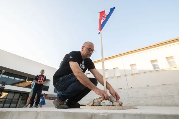 Benkovac, 06.09.2025. - U Benkovu je održan prosvjedno-performativni događaj pod nazivom "Nosi se kamen slobode" u organizaciji umjetnika i aktivista Juraja Arasa. Performans je izveden kao tiha i disciplinirana akcija. Nakon kratke obavijesti s pozornice o načinu sudjelovanja, sudionici su jedan po jedan uzeli kamen s obližnjeg parkirališta, donijeli ga do pozornice i odložili na hrpu ispod male zastave Republike Hrvatske.Na fotografiji: Juraj Aras, Foto HINA/Zvonko KUCELIN