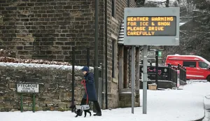 A woman walks her dog past a sign cautioning drivers about the snowy conditions in the town of Glossop, Derbyshire, northern England on January 6, 2026, after a light snow covered the region overnight. The UK's Met Office issued fresh weather warnings for January 5-6 for snow and ice for Scotland, Northern Ireland and parts of northern England and said cold weather health alerts for all English regions would remain in place until January 9. (Photo by Oli SCARFF/AFP)