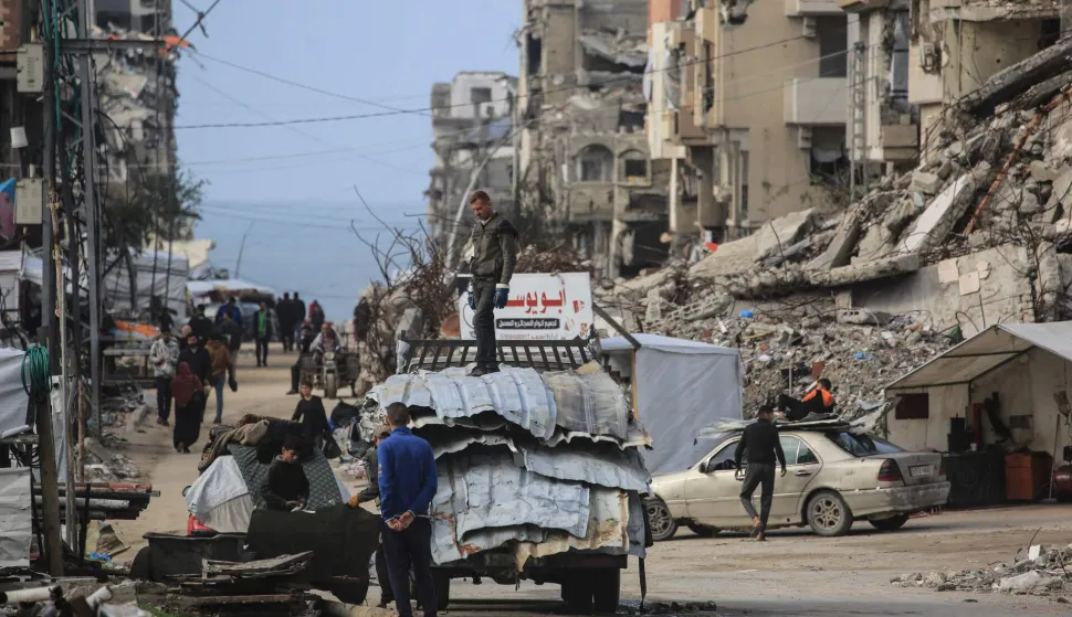 A man stands on a truck loaded with metal sheets as people make their way amid damaged buildings in Jabalia, in the northern Gaza Strip, on January 10, 2026. Since October 10, a fragile US-sponsored truce in Gaza has largely halted the fighting between Israeli forces and Hamas, but both sides have alleged frequent violations. (Photo by Bashar Taleb/AFP)