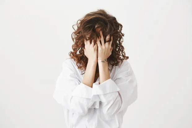 Studio portrait of tired or bothered woman hiding face in her curly hair and holding hands on it, expressing exhaustion or just being childish, standing over gray background. I am not seeing you.