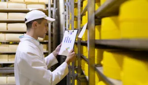 Technologist in a white robe holding graphs statistics and stands near the shelves with cheeses. Production of cheese products. racks with cheeses freepik