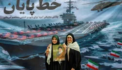 Women pose with a poster depicting Iran's supreme leader Ayatollah Ali Khamenei as they stand before a banner depicting a US Navy aircraft carrier with US-flag-wrapped coffins on the deck, during a rally marking the 47th anniversary of the 1979 Islamic revolution in Tehran on February 11, 2026. The Persian calendar date of Bahman 22 celebrates the anniversary of the resignation of the ousted shah's last prime minister and the formal assumption of power by revolutionary leader Ayatollah Ruhollah Khomeini. (Photo by AFP)