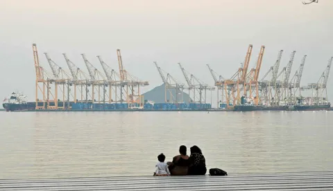 (FILES) A family sits against the backdrop of a dockyard off coast city of Fujairah, in the Strait of Hormuz in the northern Emirate on February 25, 2026. The US president sent a peace plan to Iran as he voiced optimism on March 25, 2026 at ending nearly a month of warfare, with Tehran announcing that it will let "non-hostile" oil vessels go through the crucial Strait of Hormuz. Oil prices dropped sharply and stocks in Asia rose on broader de-escalation hopes following nearly four weeks of war, as the US president appeared to be ramping up efforts to bring an end to his joint military operation with Israel. (Photo by Giuseppe CACACE/AFP)