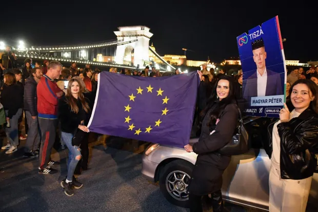 Participants display an election campaign poster of Hungary's prime minister-elect, Peter Magyar and a flag of the European Union during a spontaneous gathering to celebrate the victory of the conservative Tisza party near the Szechenyi Chain Bridge in Budapest late on April 13, 2026, one day after Hungary's general elections. Within hours of becoming Hungary's prime minister-elect, Magyar pledged on April 13 to usher in a "new era" after defeating long-time nationalist leader Orban in an election seen as a blow to hard-right populism. (Photo by Ferenc ISZA/AFP)