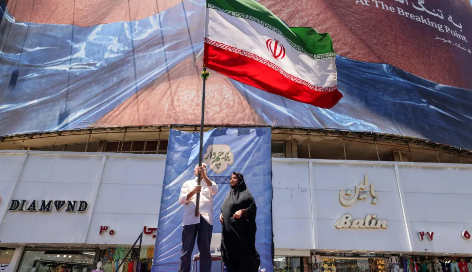 A man raises an Iranian national flag as he stands on a podium before a political billboard featuring US President Donald Trump and the Strait of Hormuz along Valiasr Square in Tehran on May 26, 2026. US forces launched overnight strikes on missile sites in Iran and boats they said were trying to lay mines in Gulf waters, sending oil prices higher on May 26, while diplomats sought a deal to end the war. Iran did not immediately confirm the reported strikes, but state media reported blasts in the southern port city of Bandar Abbas, and the Revolutionary Guards Corps said its forces had downed a US drone entering its airspace and fired at an F-35 fighter jet. (Photo by ATTA KENARE/AFP)