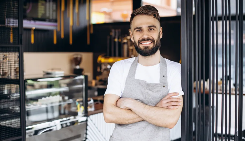 Male barista standing and smiling at a coffee houseKoliko sezonac može zaraditi?Istražili smo koliko traže radnici, a koliko nude poslodavci? Koliko hotelijeri trenutno plaćaju sobarice, recepcionere i ostalo osoblje? konobar ilustracija freepik