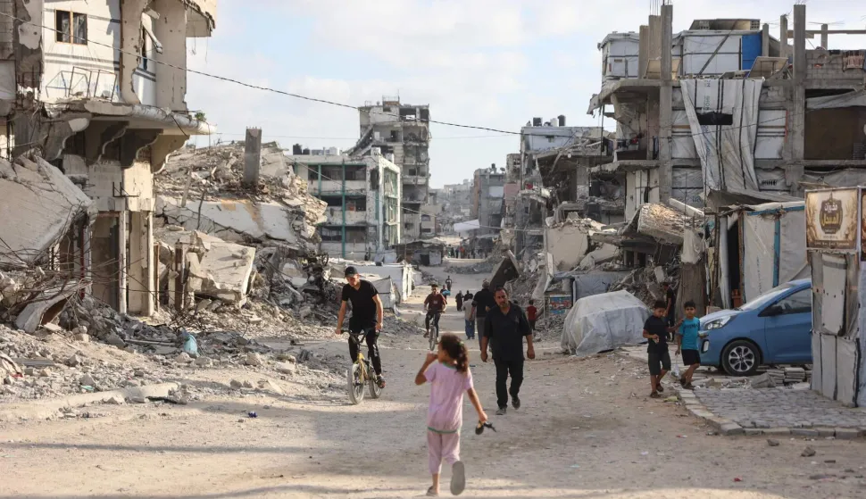 Palestinians cycle and walk along a street lined with damaged buildings near the al-Shati refugee camp, west of Gaza City, on July 1, 2026. Gaza remains gripped by daily violence, with both the Israeli military and Hamas accusing one another of violating the truce in effect since October 10. The war flattened swathes of the territory, displaced most of the population at least once, and left hundreds of thousands of people living in tents and temporary shelters. (Photo by Omar AL-QATTAA/AFP)