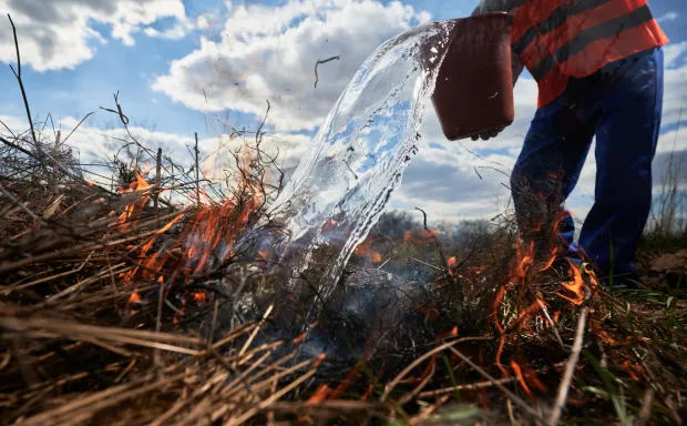 Firefighter ecologist fighting wildfire in field with cloudy sky on background. Close up of male environmentalist pouring water on burning dry grass. Natural disaster and ecology demage concept.