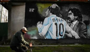 A man rides his bike past a mural depicting late Argentine football legend Diego Maradona and star forward Lionel Messi during a match at the 2010 World Cup, in Villa Fiorito, on the outskirts of Buenos Aires, on July 14, 2026. In a packed cinema, Argentinians applaud, weep and relive Diego Maradona?s goals against England in 1986 as if they were sitting in the stands. Forty years on, with the dispute over sovereignty of the Falkland Islands still ongoing and a rivalry that extends far beyond football, the July 15 semi-final for the 2026 World Cup goes beyond the pitch. (Photo by Luis ROBAYO/AFP)