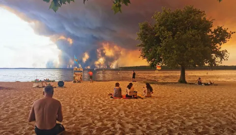 TOPSHOT - Beach goers sit on the beach of Moutchic in Lacanau, southwestern France on July 24, 2026, as clouds of smoke rise into the sky from a wildfire. The fire raging in the northern part of the Arcachon Basin in Gironde, southwest France, has swept through more than 3,400 hectares, according to the latest figures released by the prefecture. More than 20,000 residents and tourists have been evacuated from this part already devastated by flames in 2022. Scientists have long warned that climate change caused by human use of fossil fuels will make periods of drought more intense and longer-lasting, creating the ideal conditions for wildfires. (Photo by Maximilien LAMY/AFP)