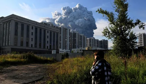 A person speaks on a mobile phone as smoke rises above residential buildings from a warehouse for a Russian e-commerce giant near Saint Petersburg after being hit by Ukrainian drones early on July 24, 2026 morning, triggering a fire that sent smoke pouring over the historic city, according to local officials. Kyiv has targeted Saint Petersburg as it steps up long-range drone strikes on Russian energy, military and logistics sites in recent weeks. Russia's defence ministry said its forces shot down some 571 drones overnight over more than a dozen regions and Crimea, the Black Sea peninsula that Russia annexed in 2014. (Photo by AFP)