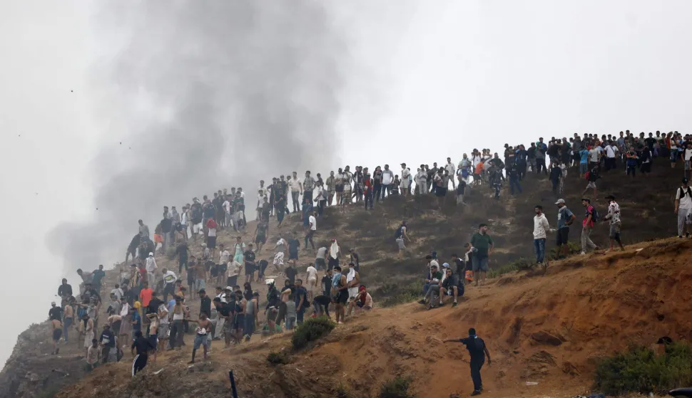 TOPSHOT - People gather on a hilltop as smoke billows from the site of clashes near Fnideq, on the Morocco-Spain border, on July 31, 2026. Thousands of migrants crossed overnight from Morocco into Spain's north Africa's Ceuta enclave, a police source said on July 31, adding that it was impossible to estimate how many people in total. A total of 18 people died while attempting to reach the Spanish enclave, the source said. (Photo by Abdel Majid BZIOUAT/AFP)