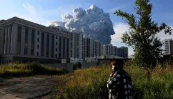 A person speaks on a mobile phone as smoke rises above residential buildings from a warehouse for a Russian e-commerce giant near Saint Petersburg after being hit by Ukrainian drones early on July 24, 2026 morning, triggering a fire that sent smoke pouring over the historic city, according to local officials. Kyiv has targeted Saint Petersburg as it steps up long-range drone strikes on Russian energy, military and logistics sites in recent weeks. Russia's defence ministry said its forces shot down some 571 drones overnight over more than a dozen regions and Crimea, the Black Sea peninsula that Russia annexed in 2014. (Photo by AFP)