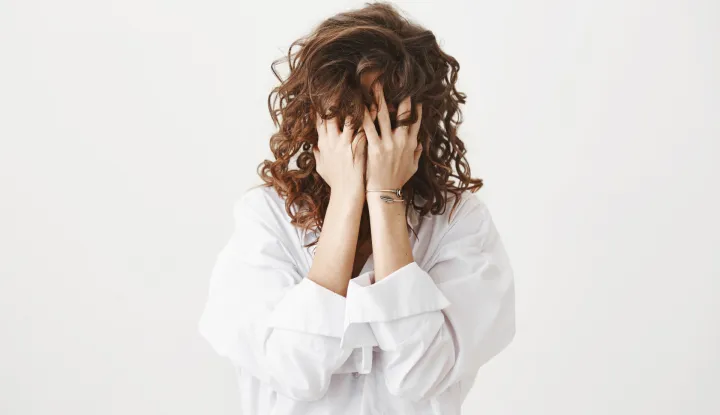 Studio portrait of tired or bothered woman hiding face in her curly hair and holding hands on it, expressing exhaustion or just being childish, standing over gray background. I am not seeing you.
