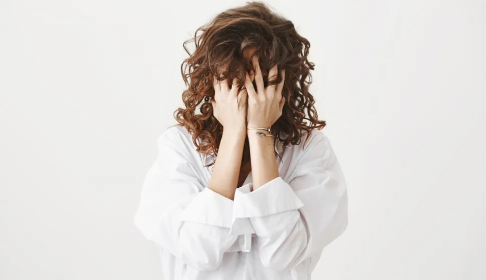 Studio portrait of tired or bothered woman hiding face in her curly hair and holding hands on it, expressing exhaustion or just being childish, standing over gray background. I am not seeing you.
