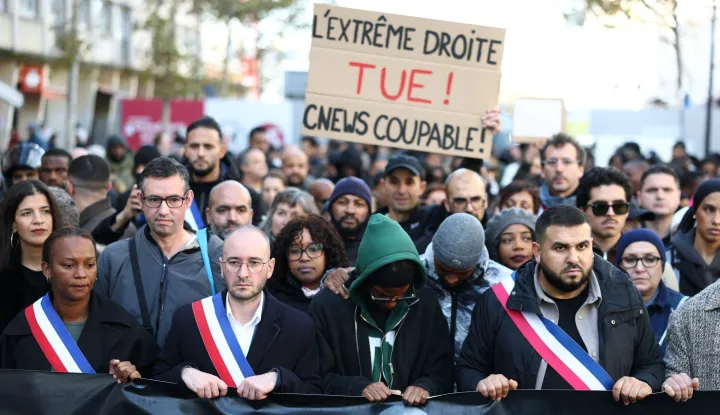 TOPSHOT - Attendees and officials including mayor of Aubervilliers Sofienne Karroumi (R) with an attendee in the background holding a placard reading "the far right kills ! CNEWS guilty !" referencing French TV channel, take part in a silent march in tribute to Pengo Bossio Wangi, a man killed in a shooting by an attacker on October 23, a march organised by his MMA club near the town hall of Aubervilliers, north of Paris, on October 25, 2026. A man suspected of a gun attack in the Paris suburbs that killed Pengo Bossio Wangi and wounded five other persons has left custody and been placed in a psychiatric unit for observation, French anti-terror prosecutors said on October 25, 2026. The 39-year-old suspect, partly identified as Kevin T., was arrested on October 23, in possession of a submachine gun and tear-gas grenades in his car, shortly after the victims were shot as a vehicle drove through the working-class town of Trappes and neighbouring La Verriere. (Photo by Sameer AL-DOUMY/AFP)