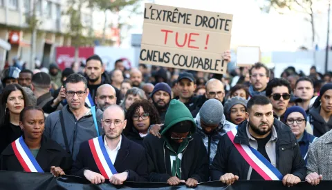 TOPSHOT - Attendees and officials including mayor of Aubervilliers Sofienne Karroumi (R) with an attendee in the background holding a placard reading "the far right kills ! CNEWS guilty !" referencing French TV channel, take part in a silent march in tribute to Pengo Bossio Wangi, a man killed in a shooting by an attacker on October 23, a march organised by his MMA club near the town hall of Aubervilliers, north of Paris, on October 25, 2026. A man suspected of a gun attack in the Paris suburbs that killed Pengo Bossio Wangi and wounded five other persons has left custody and been placed in a psychiatric unit for observation, French anti-terror prosecutors said on October 25, 2026. The 39-year-old suspect, partly identified as Kevin T., was arrested on October 23, in possession of a submachine gun and tear-gas grenades in his car, shortly after the victims were shot as a vehicle drove through the working-class town of Trappes and neighbouring La Verriere. (Photo by Sameer AL-DOUMY/AFP)